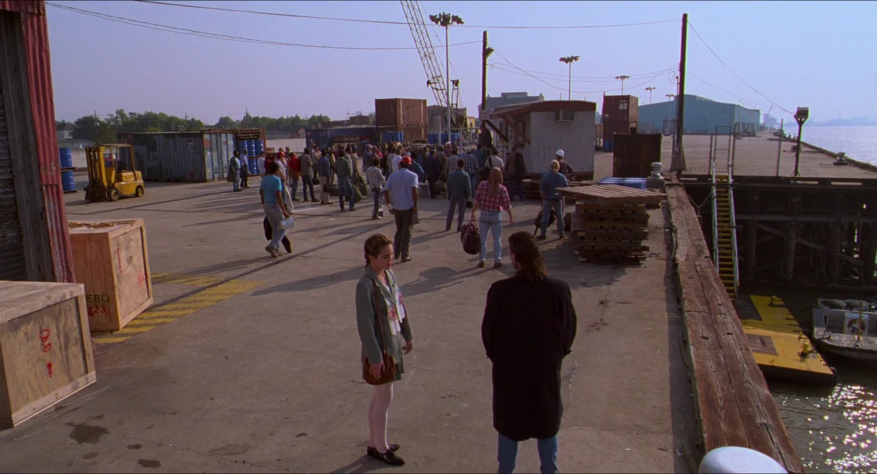 Hard Target (1993) movie still: wide — A bustling shipping pier with scattered wooden crates and industrial containers under bright sky. A woman…