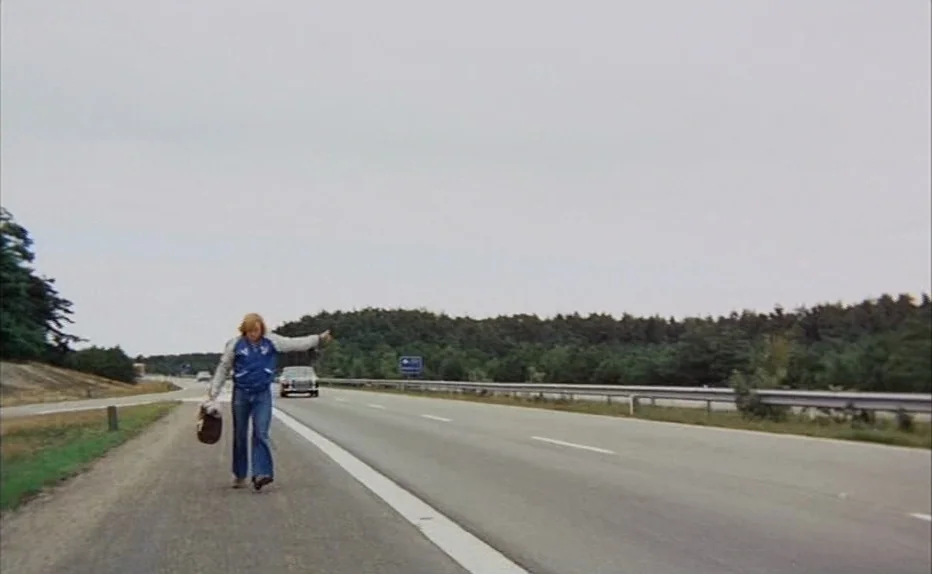 Turkish Delight (1973) movie still: wide — A wide, overcast highway shoulder stretches beside a dense line of pine trees. A young man walks toward…