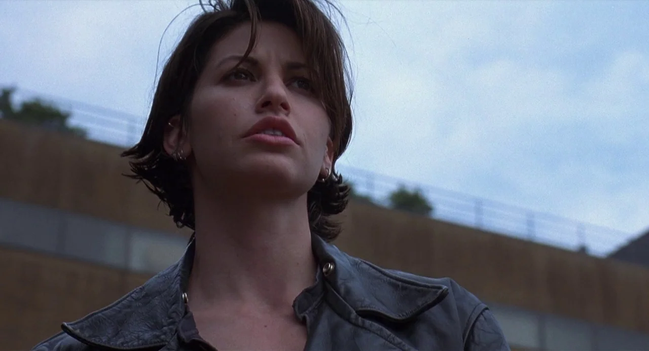 Bound (1996) movie still: closeup — A woman stands in front of a tan brick building under a cloudy sky. She looks up into the air while wearing a…