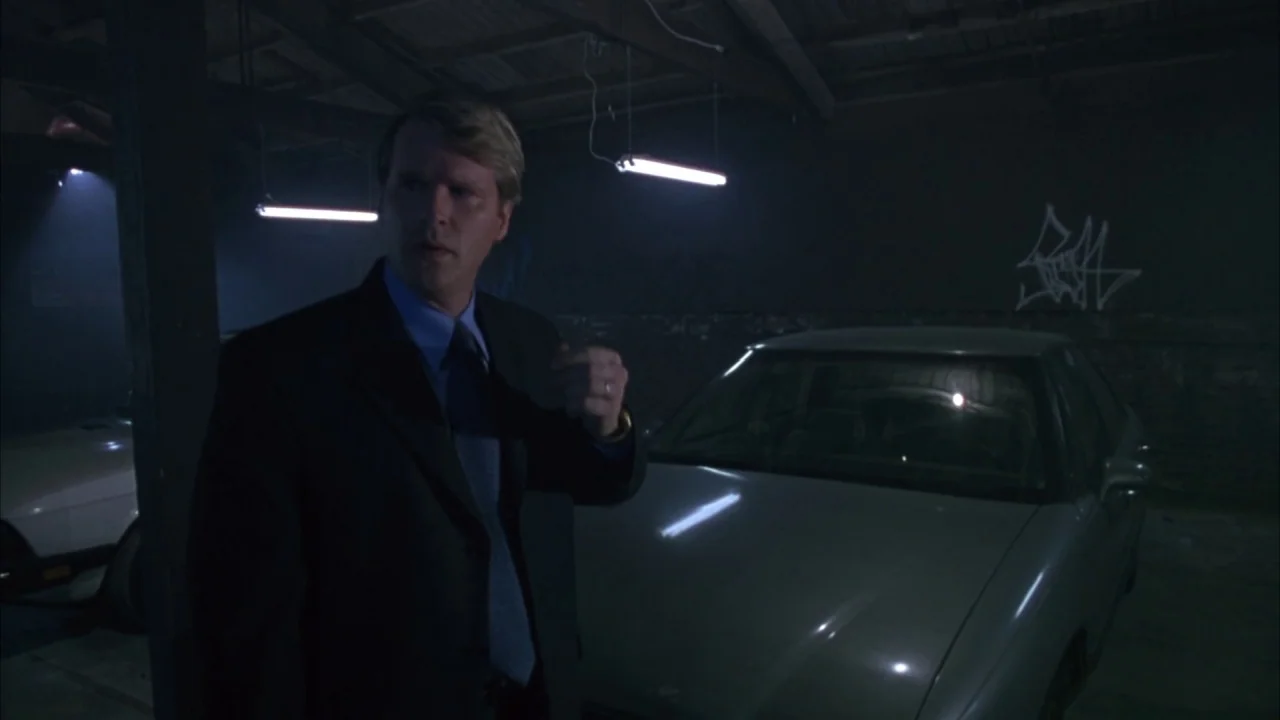 Saw movie still: medium — A dimly lit industrial parking structure with exposed rafters and graffiti on brick walls. A man in a dark business suit…