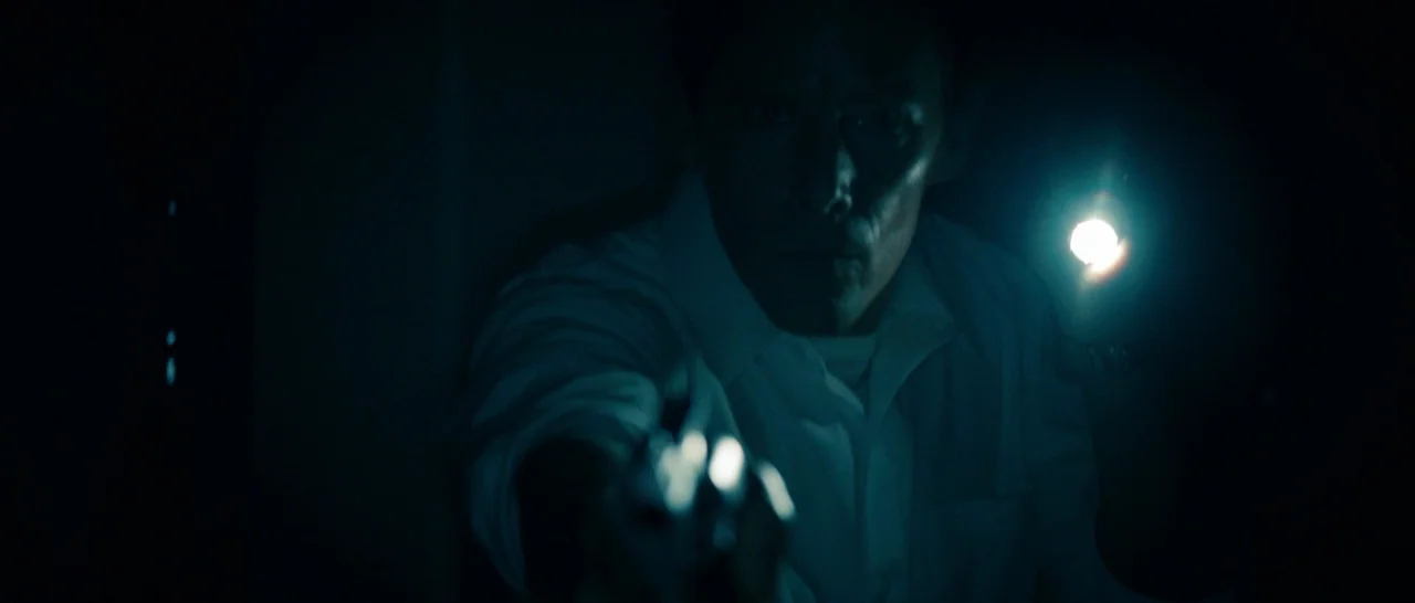 The Purge (2013) movie still: closeup — A man stands in an dark, undifferentiated interior space with deep black shadows. He holds a bright…