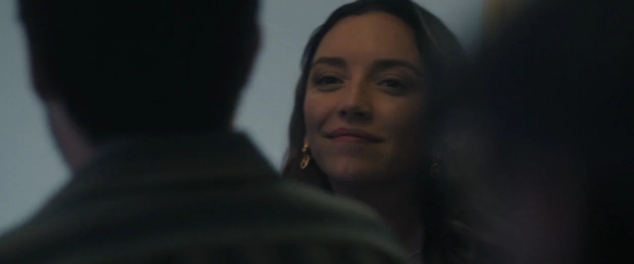 Sign Here (2017) movie still: closeup — An interior space with a neutral wall in the background. A woman looks at a man whose blurry head and…