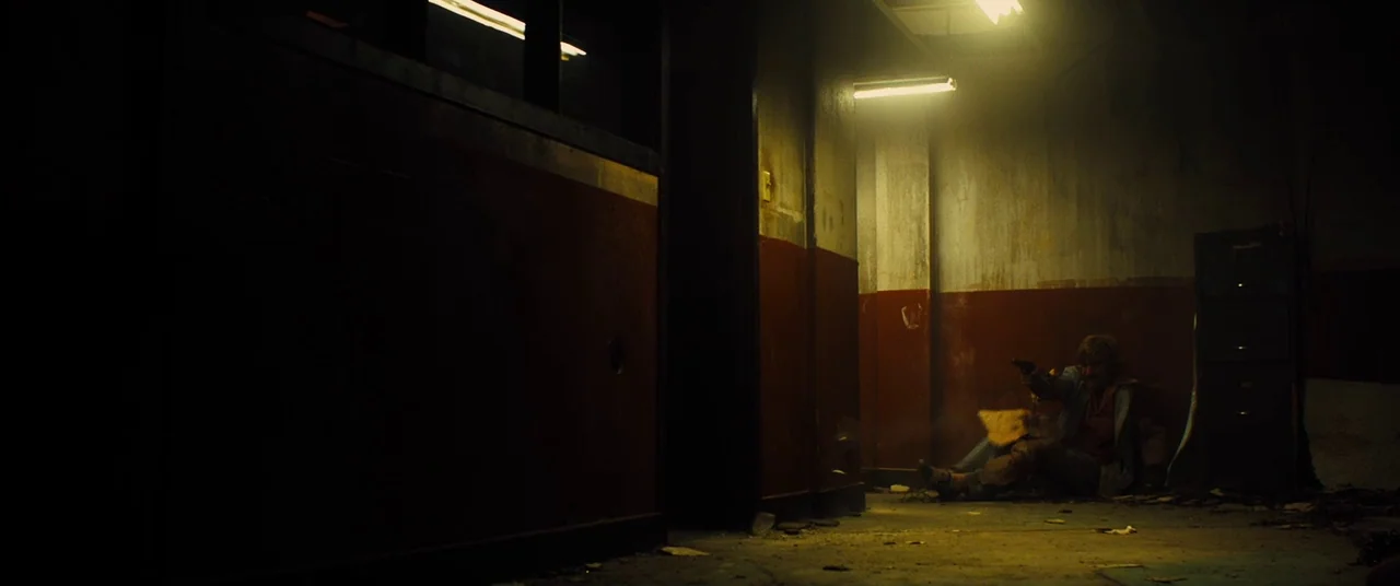 Free Fire (2016) movie still: wide — A dimly lit industrial hallway features paint-chipped walls and exposed concrete floors. A man sits against the…