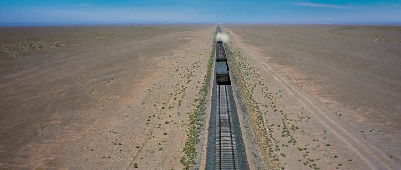 The Good The Bad The Weird (2008) movie still: establishing — An arid, flat desert landscape stretches to the horizon under a clear blue sky. A…