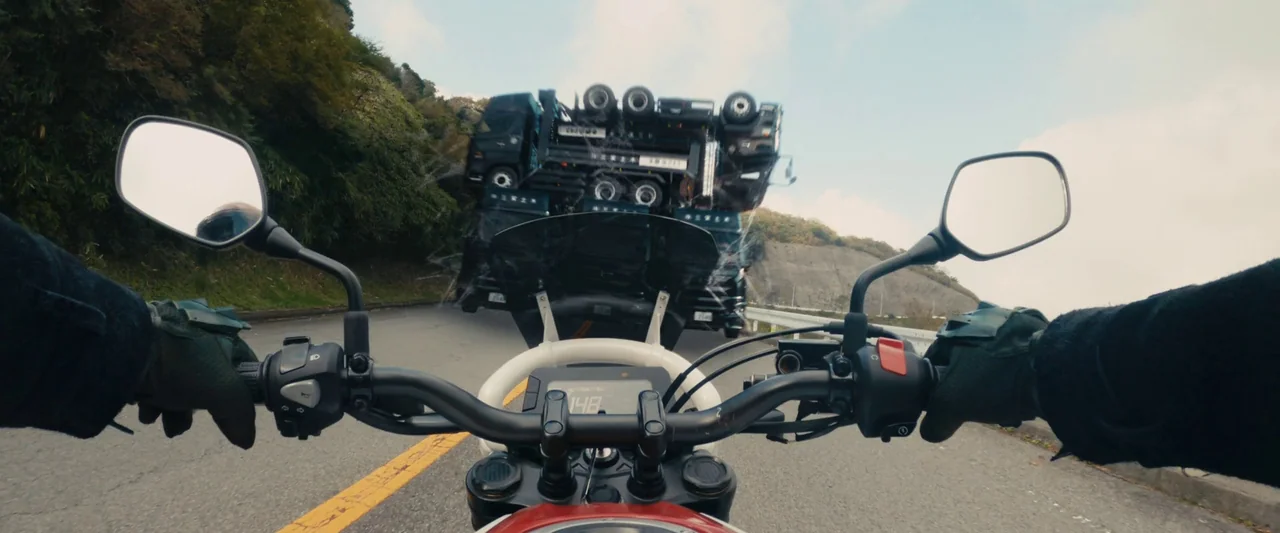 Shin Kamen Rider (2023) movie still: closeup — A view from the handlebars of a motorcycle on a winding mountain road. A large car-hauling truck…