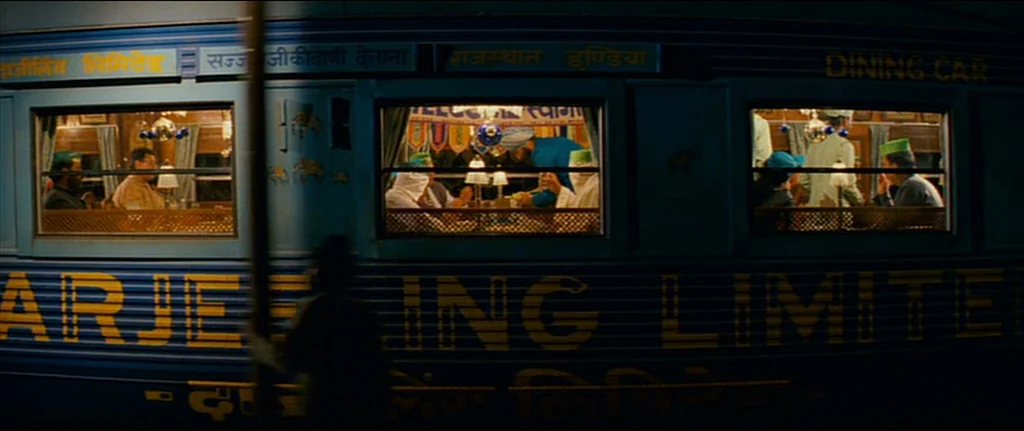 The Darjeeling Limited (2007) movie still: wide — A dark blue passenger train moves horizontally past the camera at night. Interior dining car…
