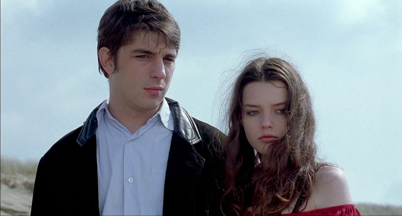Fat Girl (2001) movie still: medium — Two individuals stand in an open outdoor area under a clear, pale blue sky. The pair stands together while…