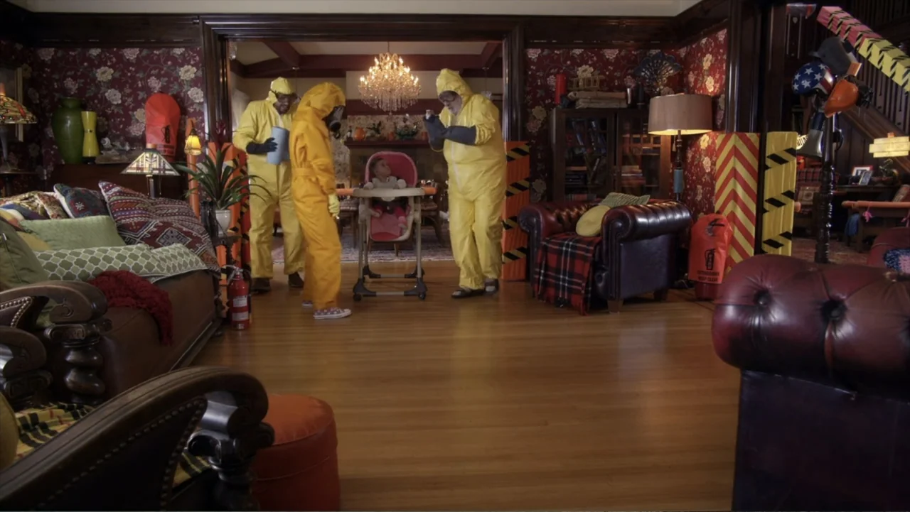 Jinxed (2013) movie still: wide — A living room with wood-paneled walls and floral wallpaper contains a dining area setup. Three men wearing yellow…