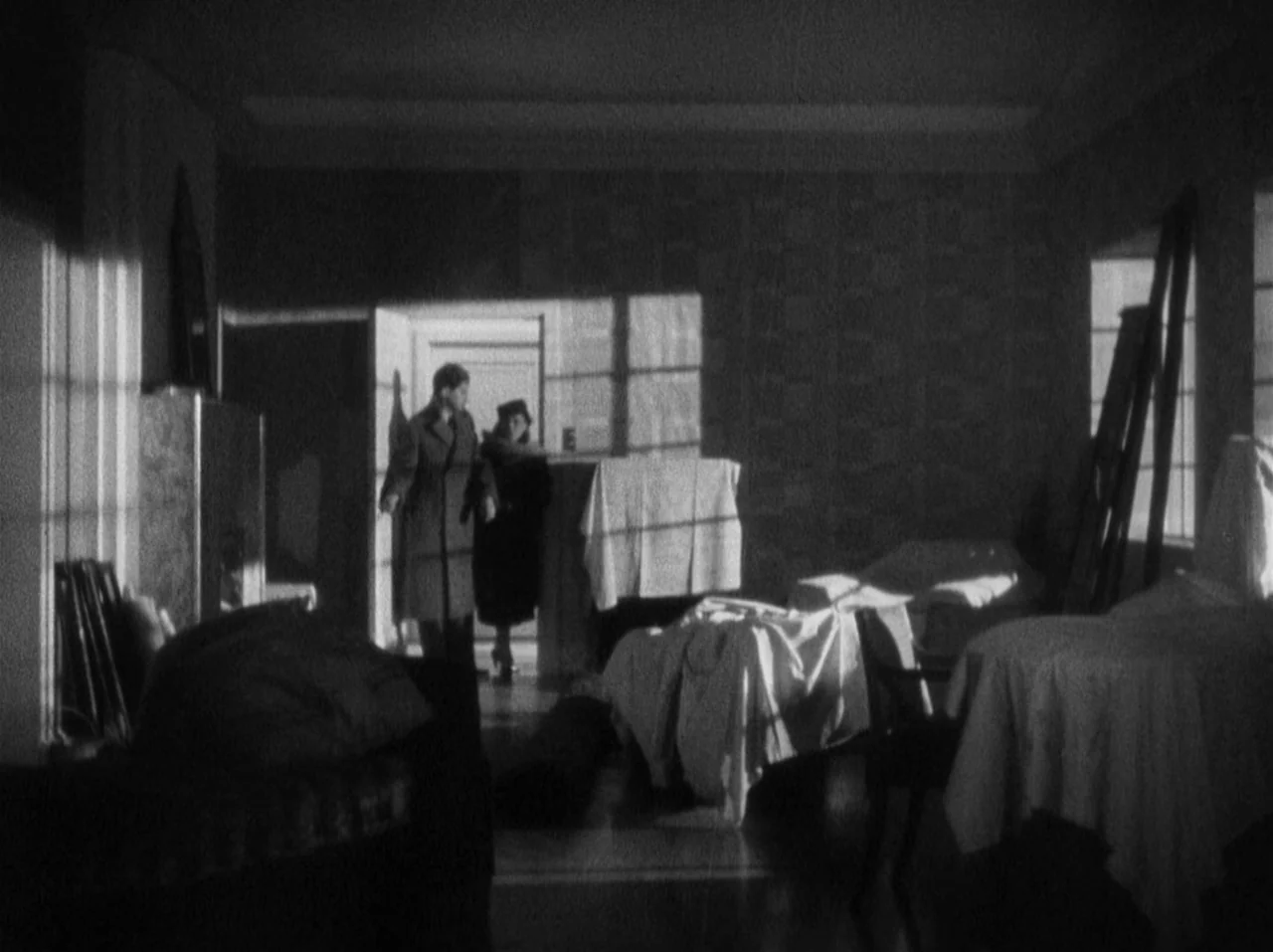 The 39 Steps (1935) movie still: wide — An empty room contains furniture items covered in large white dust sheets. A man and a woman walk through…