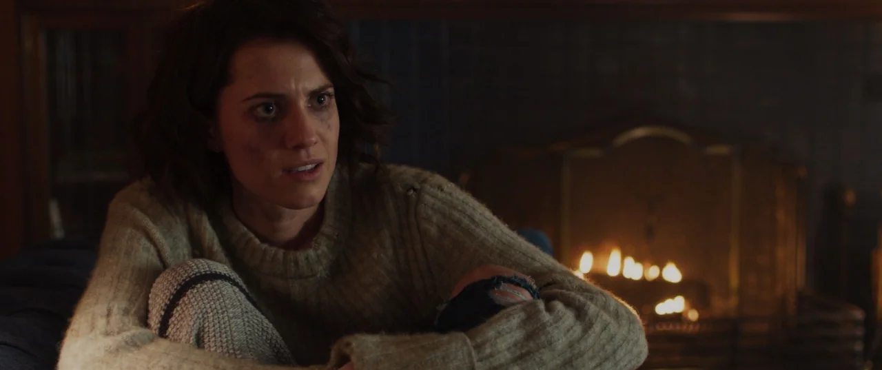 The Perfection (2018) movie still: medium — A woman sits in a dimly lit room with a burning fireplace behind her. She wears a worn beige sweater and…