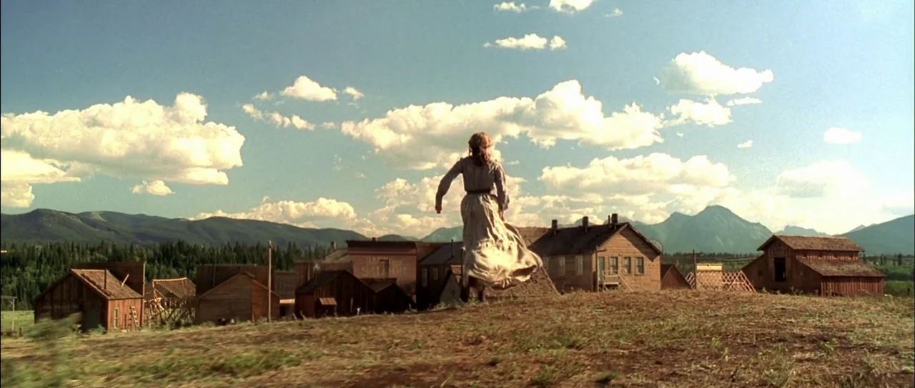 Open Range (2003) movie still: wide — A remote western frontier settlement features wooden buildings nestled beneath a mountain range. A woman…