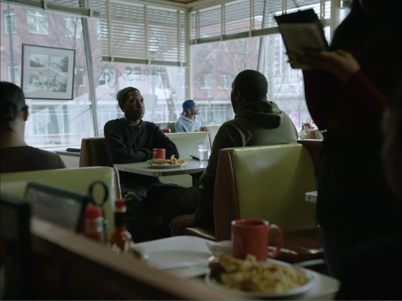 As We Speak: Rap Music on Trial (2024) movie still: medium — An interior view of a classic diner with green vinyl booths and large windows. Two men…