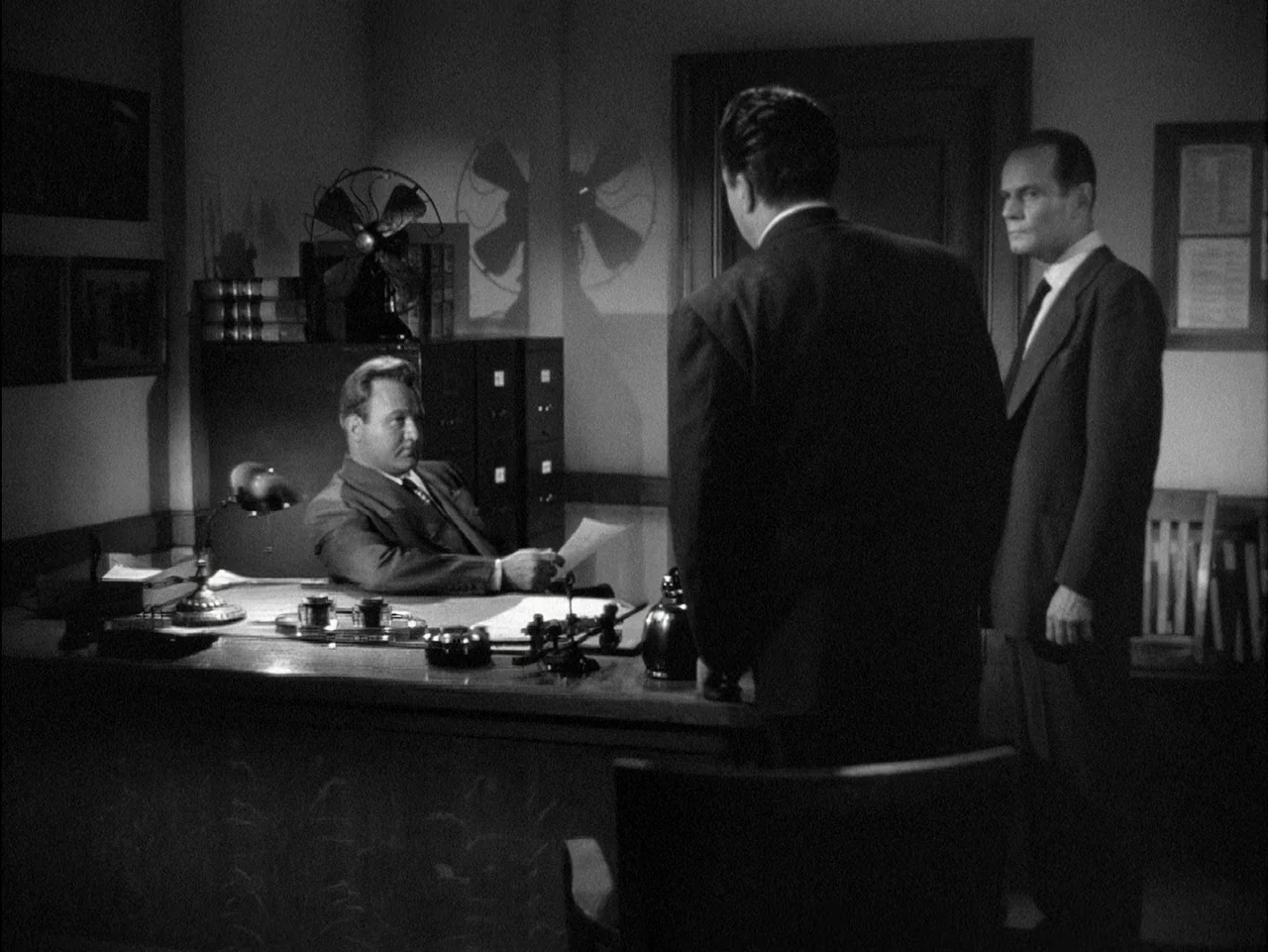 D.O.A. (1949) movie still: medium — A dimly lit 1940s interior space features a metal desk and filing cabinets. Two men stand facing a third man…