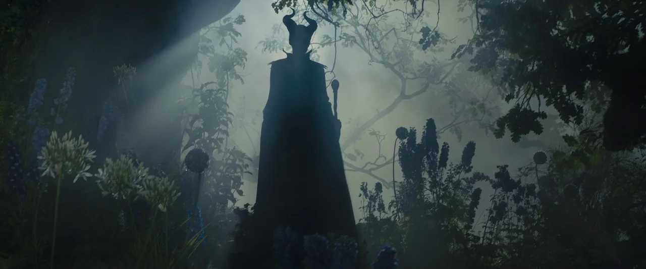 Maleficent (2014) movie still: fullbody — A dense forest clearing is filled with fog and tall blue-flowering plants. A horned silhouette stands in…