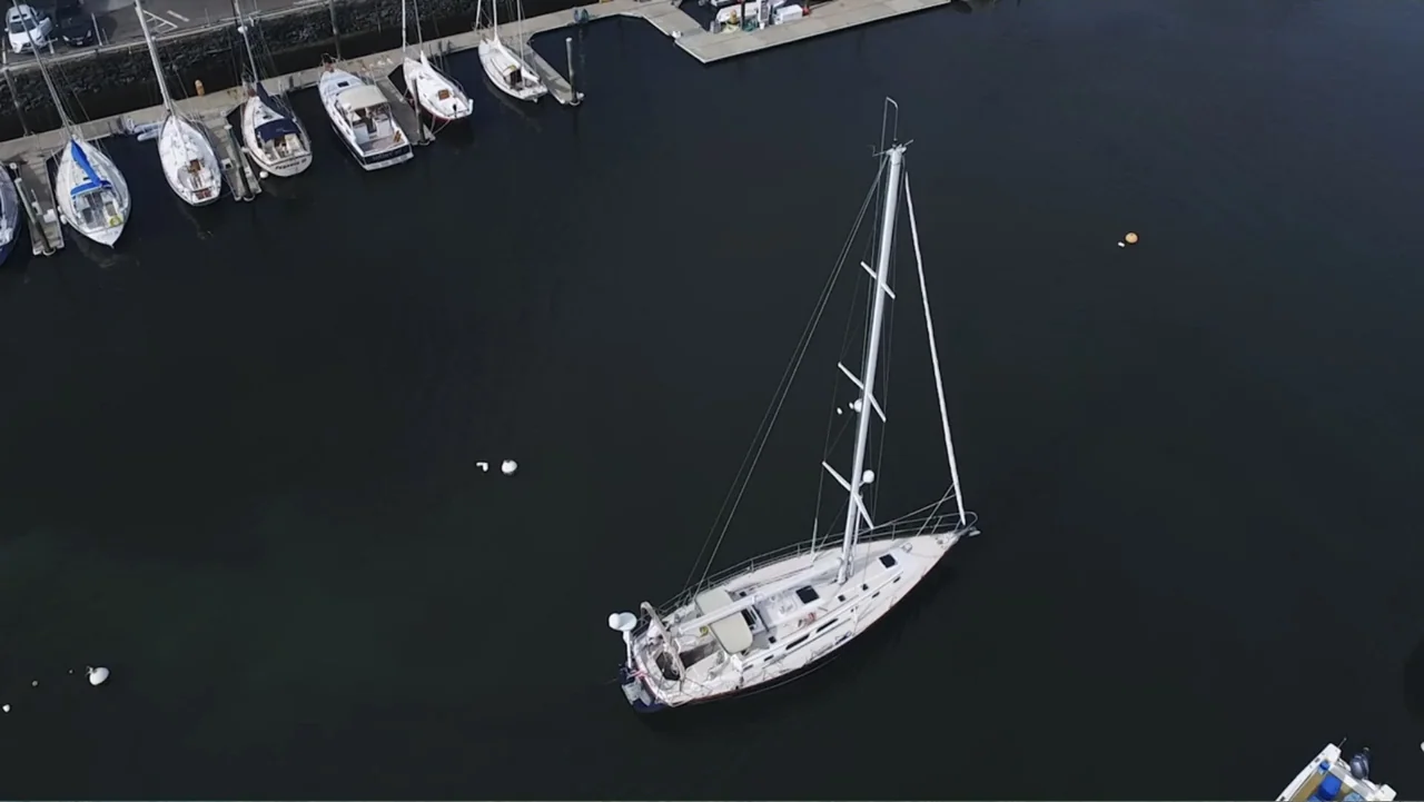 Capsized: Blood in the Water (2019) movie still: wide — A dark water basin holds a single sailboat sailing away from several docked vessels. The…