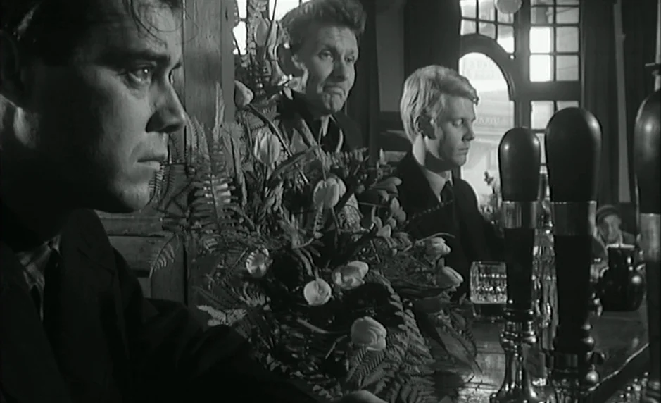 The Servant (1963) movie still: medium — A dimly lit 1960s British pub interior features a floral arrangement on the bar. Three men sit along the…