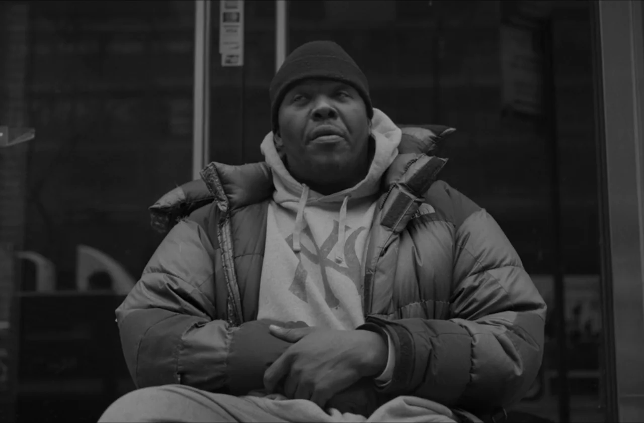 The Kitchen (2012) movie still: medium — A man sits in front of a glass storefront with dark reflective windows. He wears a heavy puffer jacket and…