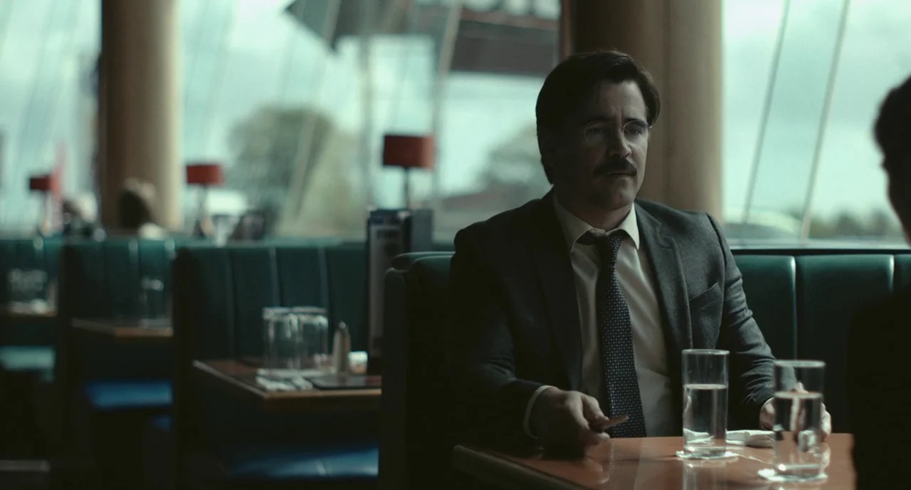 The Lobster (2015) movie still: medium — A man sits in a booth at a diner with green upholstered benches. He occupies the frame while conversing…