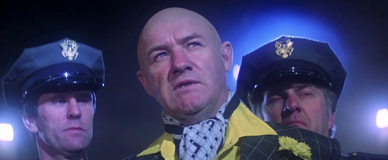 Superman (1978) movie still: medium — A bald man in a yellow shirt and plaid jacket stands between two police officers. They are positioned outdoors…