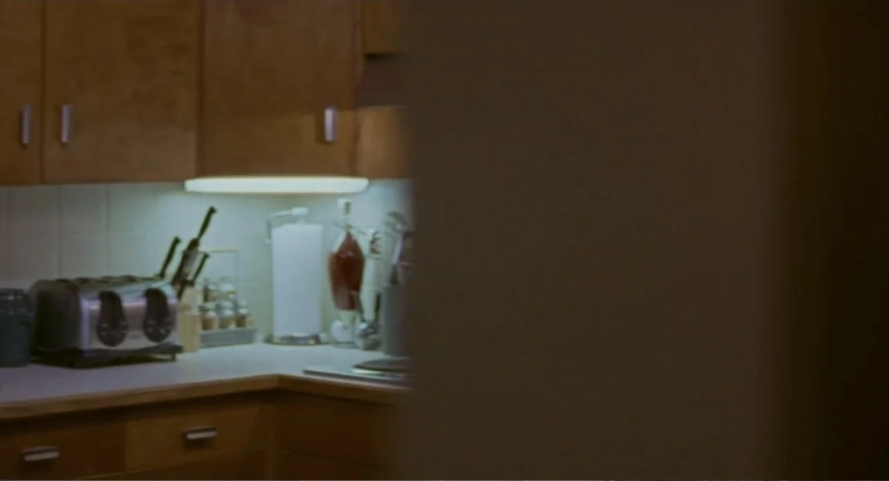 Forty Shades of Blue (2005) movie still: medium — A residential kitchen features wooden cabinets and a countertop cluttered with appliances. The…