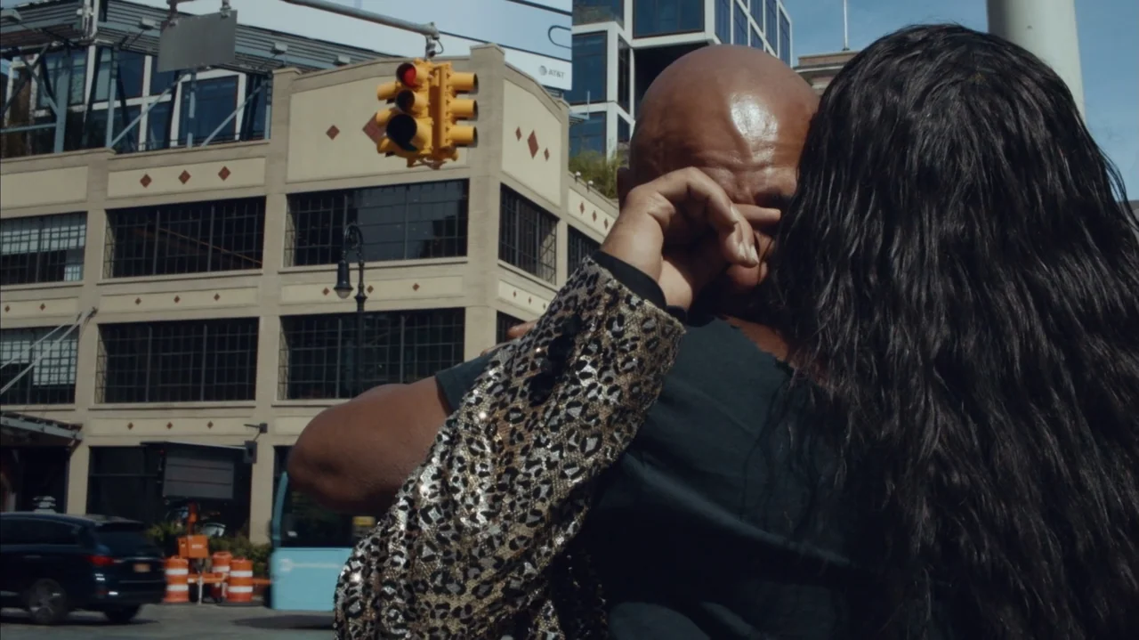 The Stroll (2023) movie still: medium — A bustling urban intersection features modern buildings and a bright yellow traffic light. Two people stand…