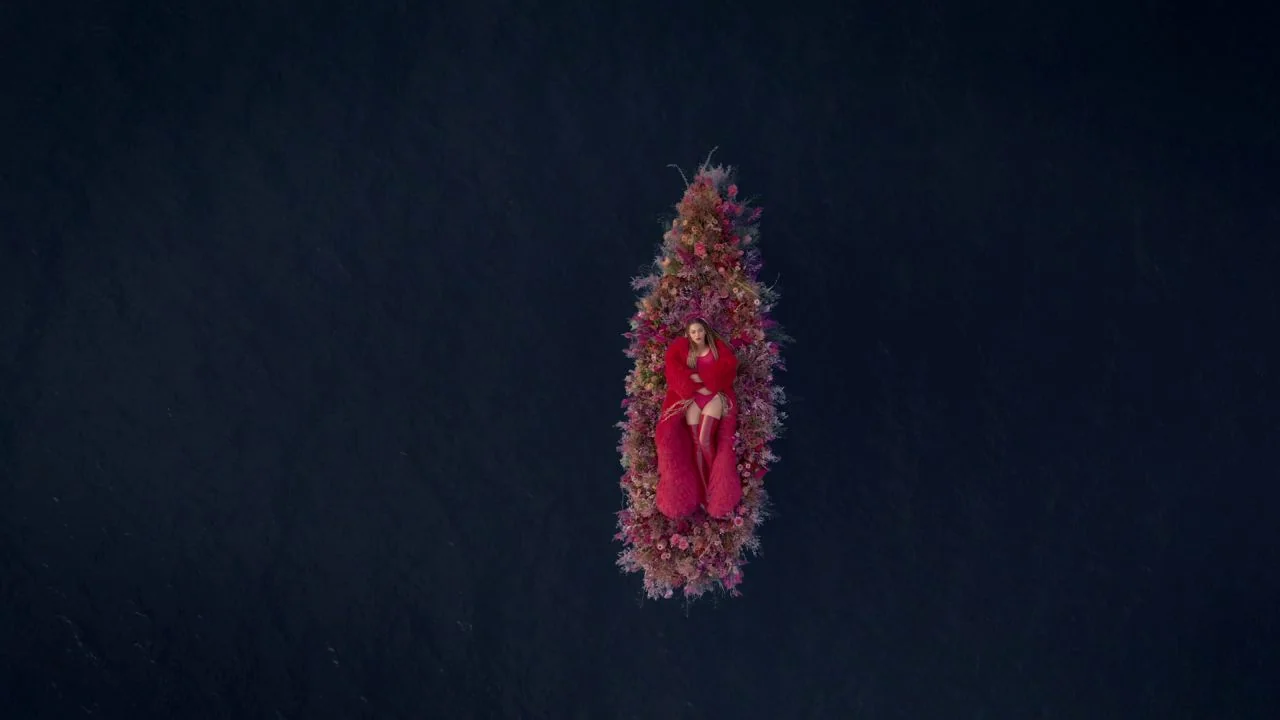 Black is King (2020) movie still: wide — A woman lies on a dense rectangular raft of pink and purple flowers. The floral structure floats in the…