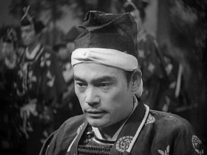 The Men Who Tread on the Tigers Tail (1945) movie still: closeup — A man in traditional Japanese headgear stands before a group of figures in…