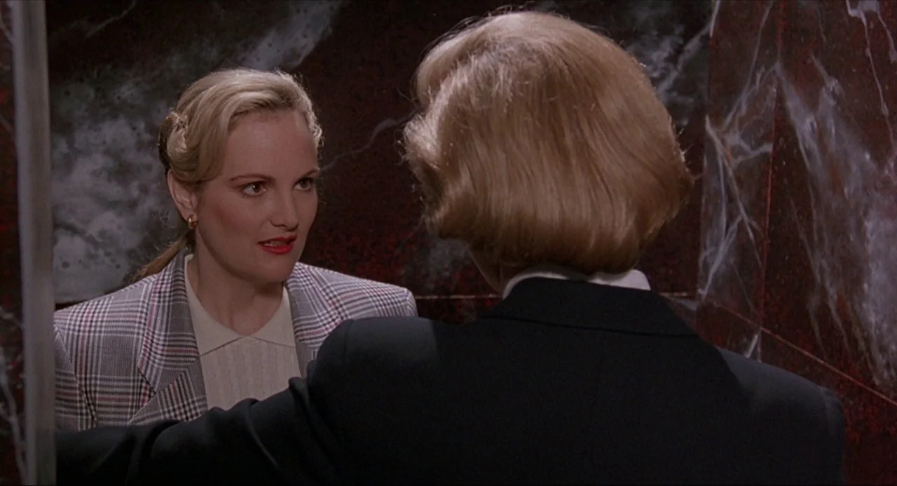 Serial Mom (1994) movie still: medium — A restroom interior with dark red marble wall cladding. One woman stands facing another woman who is…