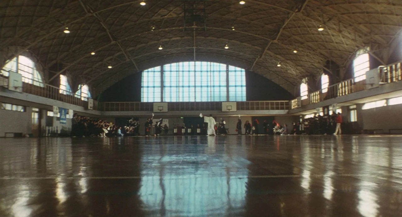 School in the Crosshairs (1981) movie still: wide — A large indoor sports facility with a polished wooden floor and high vaulted ceilings. Several…