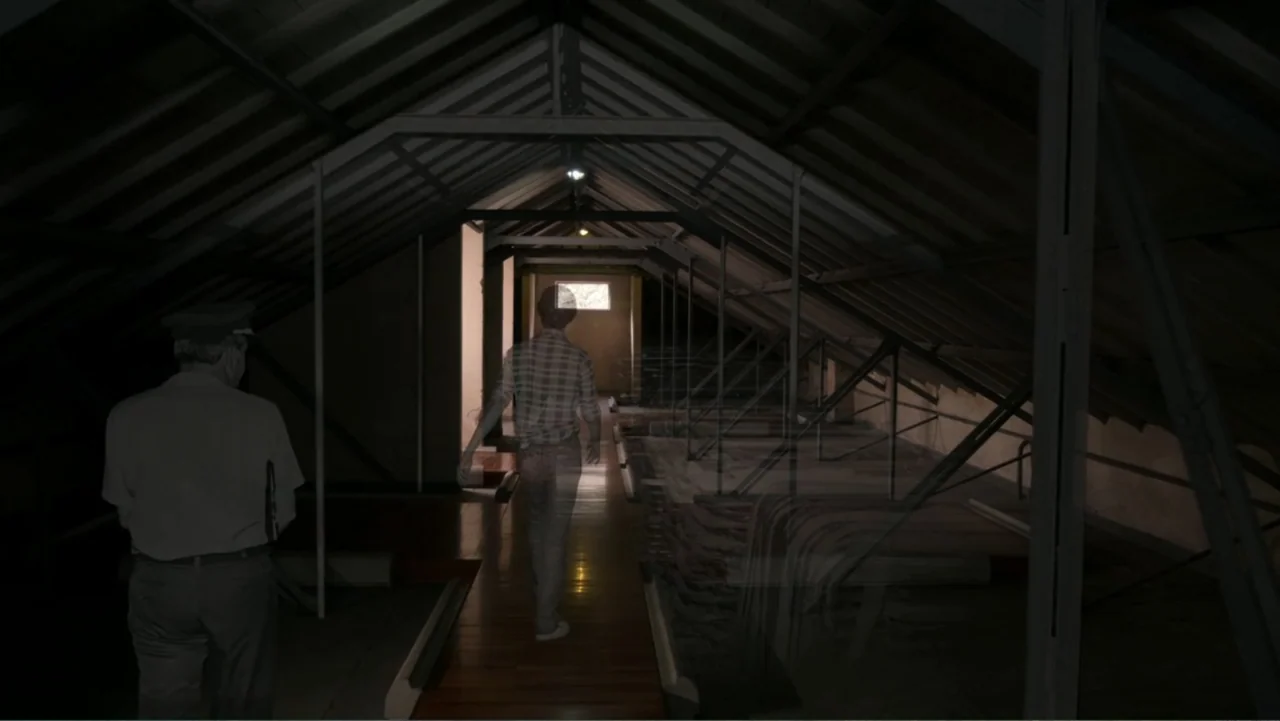 Transfers (2024) movie still: medium — A long, dark attic space features exposed wooden rafters and a central walkway. Two figures walk away from…