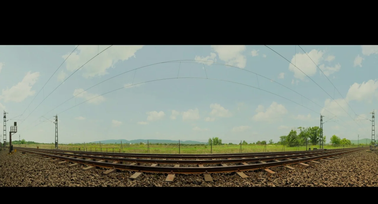 Gemini Man (2019) movie still: establishing — Curving railroad tracks span across a grassy field under a wide, cloud-speckled sky. Metal utility…