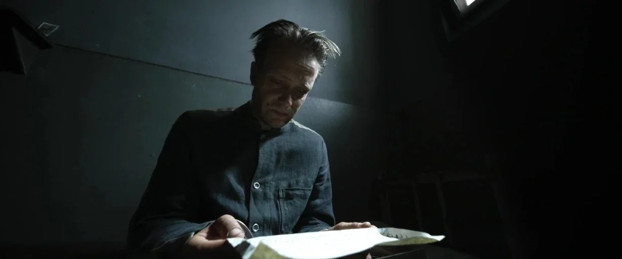 A Hidden Life (2019) movie still: medium — A dark prison cell features a single, small, high-placed window. A thin man in a grey shirt sits in the…