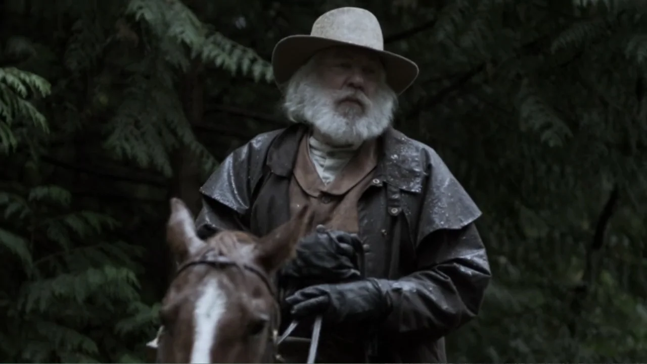 Dawn Rider (2012) movie still: medium — A dense grove of evergreen trees serves as a dark backdrop for a rider. An older man with a white beard…