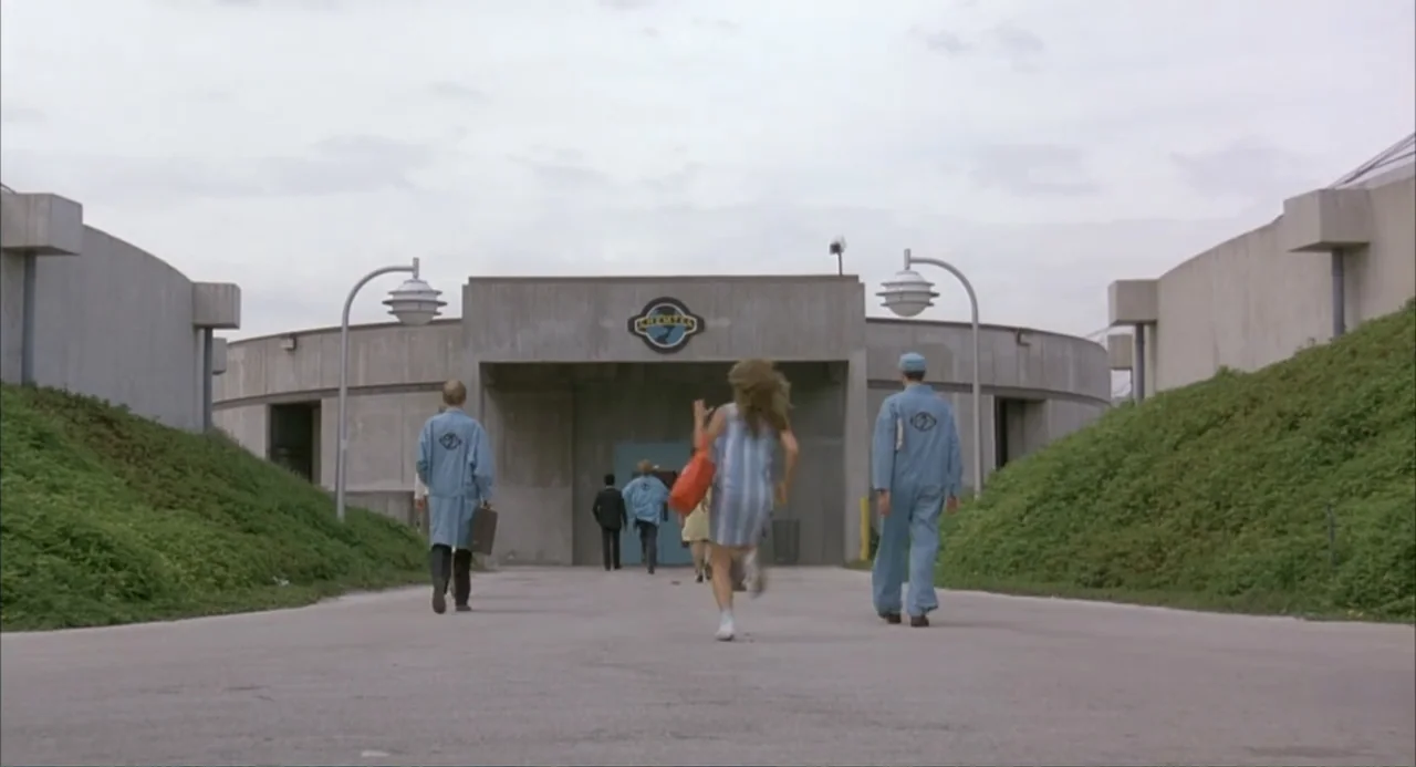 Making Mr. Right (1987) movie still: wide — A group of people approaches a brutalist concrete facility under an overcast sky. A woman in a striped…