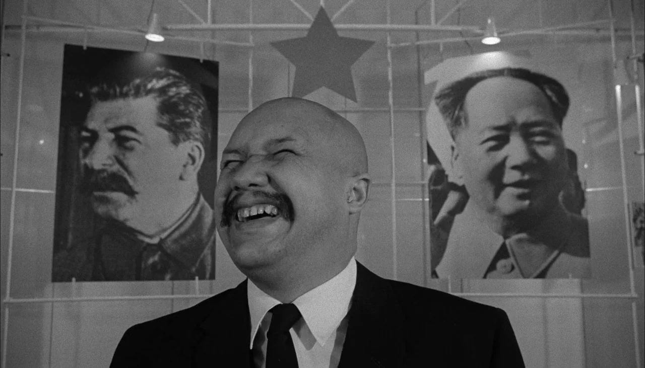 The Manchurian Candidate (1962) movie still: medium — A man stands in front of a white wall decorated with propaganda portraits and a star. The…
