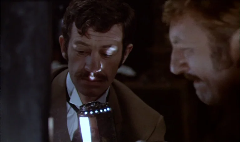 The Thief of Paris (1967) movie still: medium — A dimly lit room holds two men in 1970s attire examining a glowing object. They lean in closely to…