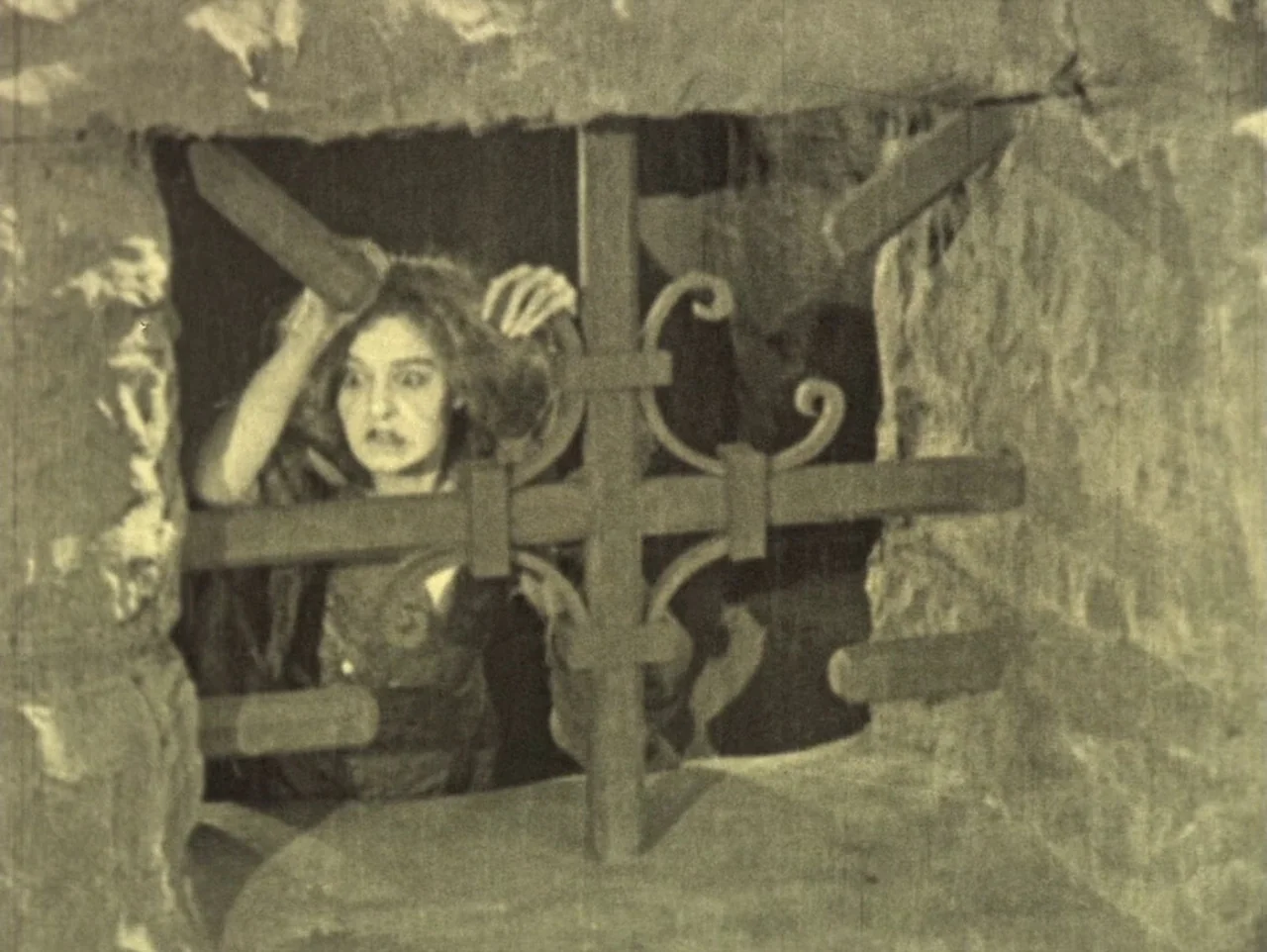 The Hunchback of Notre Dame (1923) movie still: medium — A stone-walled dungeon chamber features a dark iron grate covering an opening. A woman with…