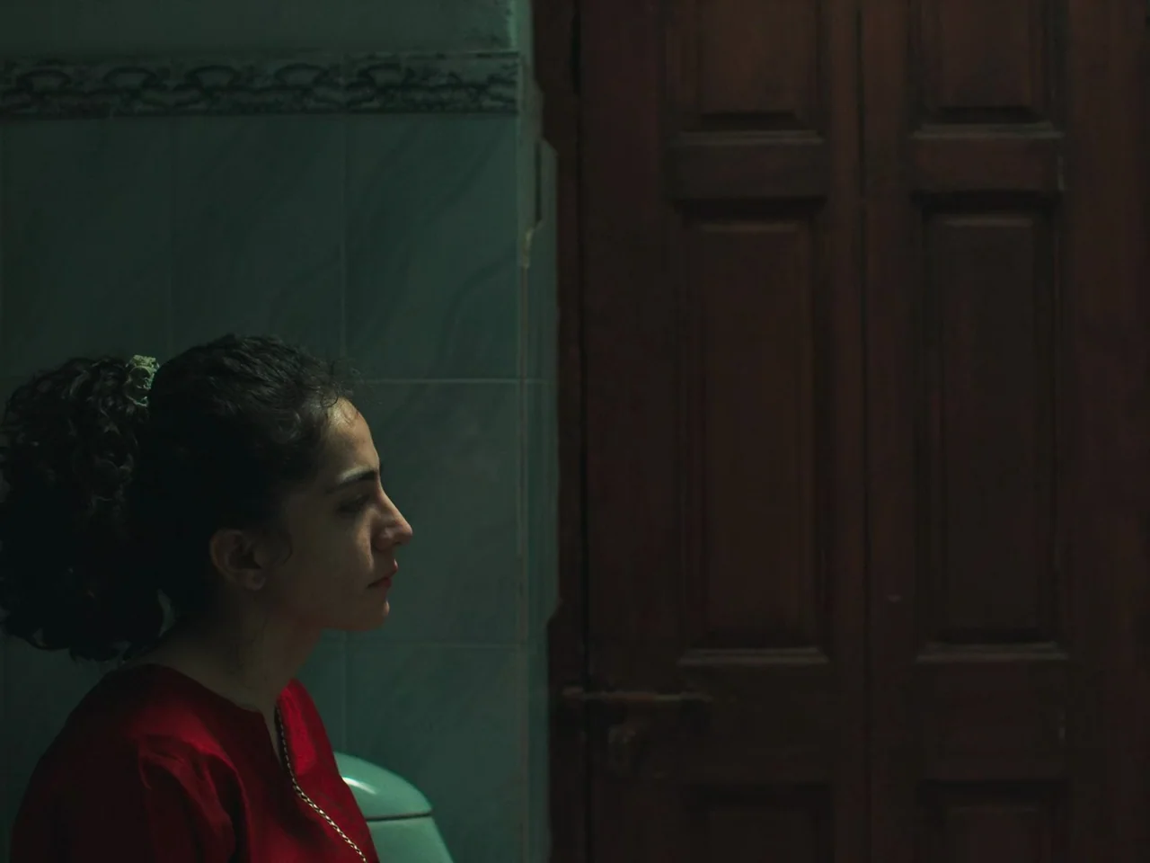 Joyland (2022) movie still: medium — A dimly lit room with light teal tiled walls and a dark wooden door. A woman in a red shirt sits in profile…