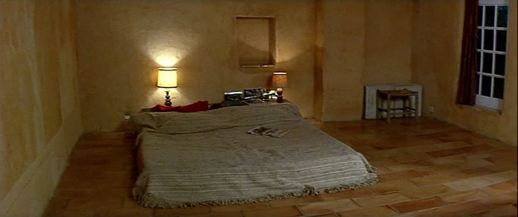 Love Unto Death (1984) movie still: wide — A spare room with terracotta tiled floors and plastered walls lit by two lamps. A low bed sits in the…