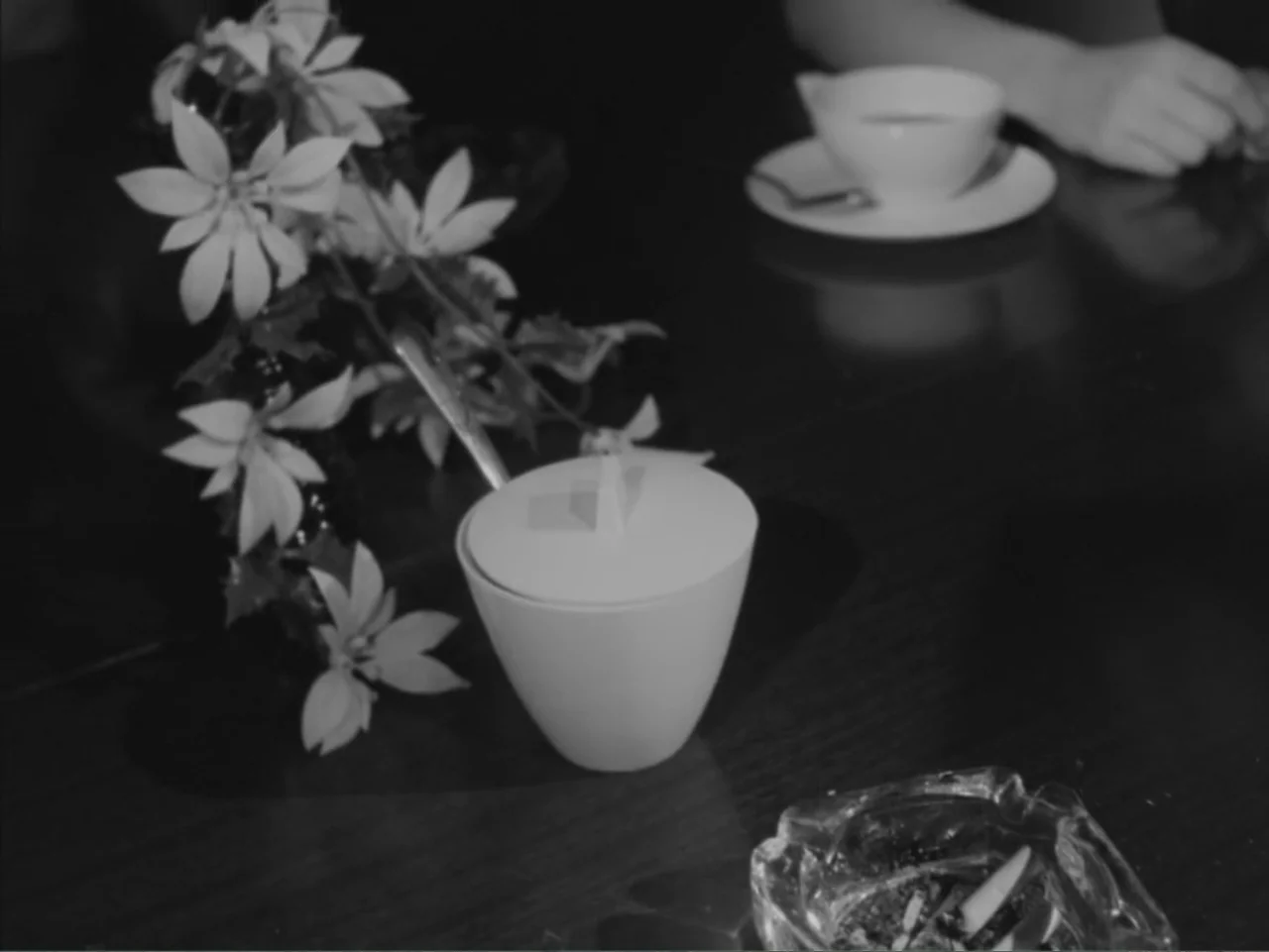 Chained Girls (1965) movie still: closeup — A dark wooden surface holds a sugar bowl, a flower arrangement, and an ashtray. Human hands rest near a…