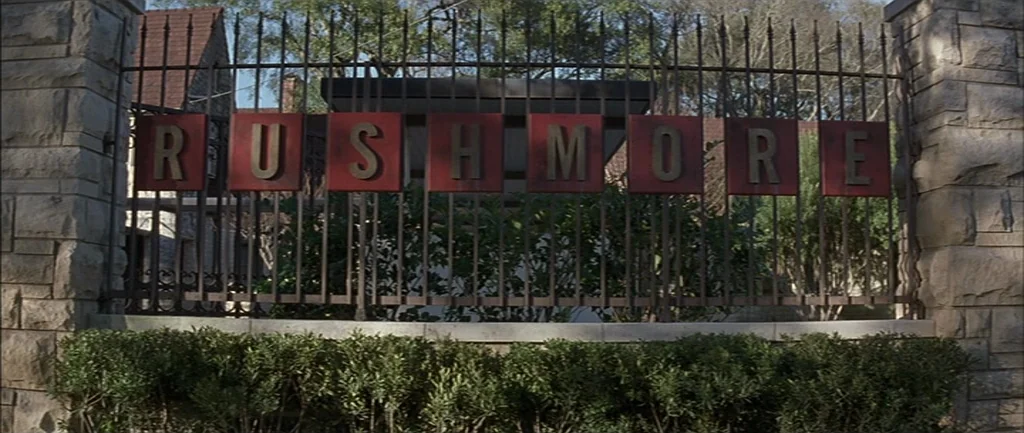 Rushmore (1998) movie still: establishing — An iron gate with individual red placards spelling out the name Rushmore sits outdoors. The gate is…