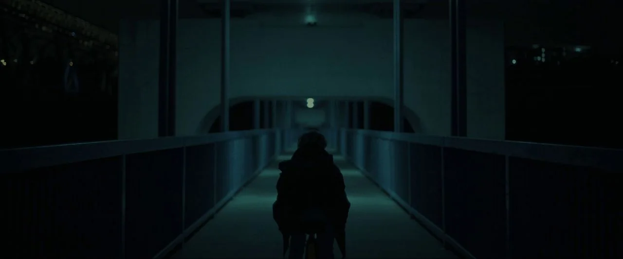 Come True (2020) movie still: fullbody — A dimly lit pedestrian bridge stretches into the darkness at night. A young person on a bicycle moves away…