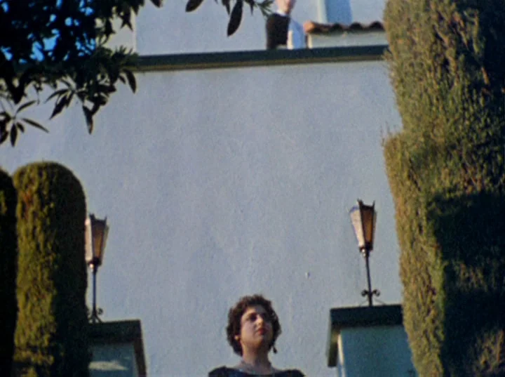 Puce Moment (1949) movie still: medium — A white stucco wall rises behind a woman standing on a terrace. A man in a suit is visible on a balcony…