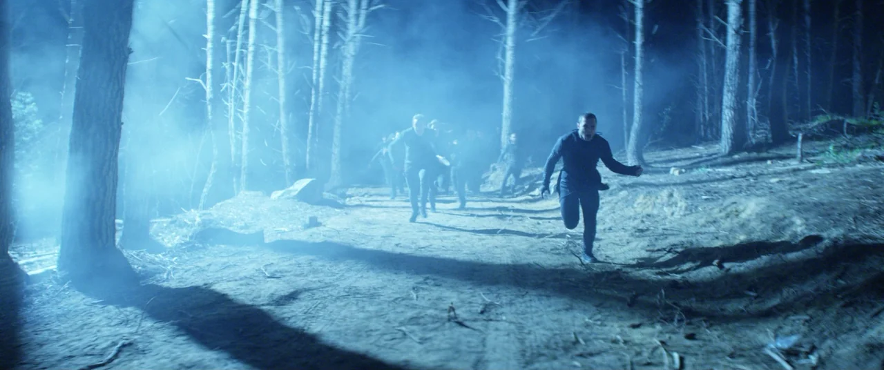 The Empty Man (2020) movie still: wide — A group of men runs through a dense, foggy nighttime pine stand. Tall thin trees flank a dirt trail that…