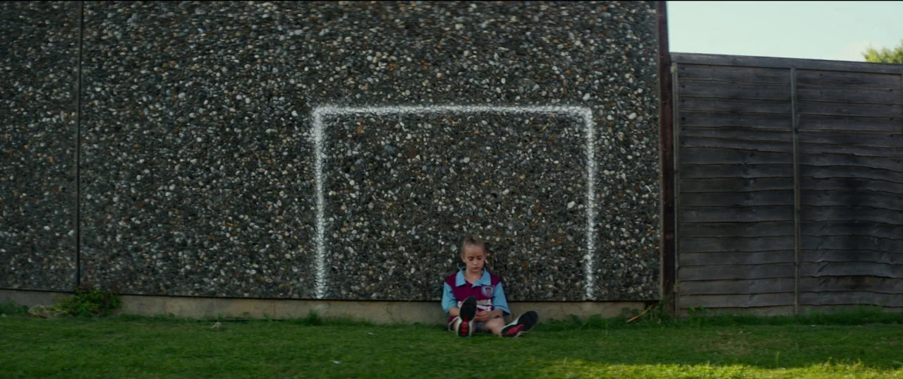 Scrapper (2023) movie still: wide — A gravel-dashed wall features a painted white rectangle representing a goal. A young child sits on the grass in…