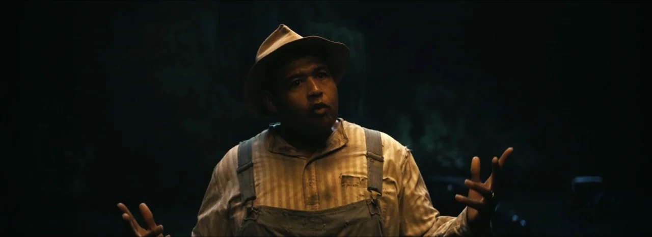 Sinners (2025) movie still: medium — A dark outdoor space at night shows the upper torso of a man. The man wears old-fashioned work clothes and…
