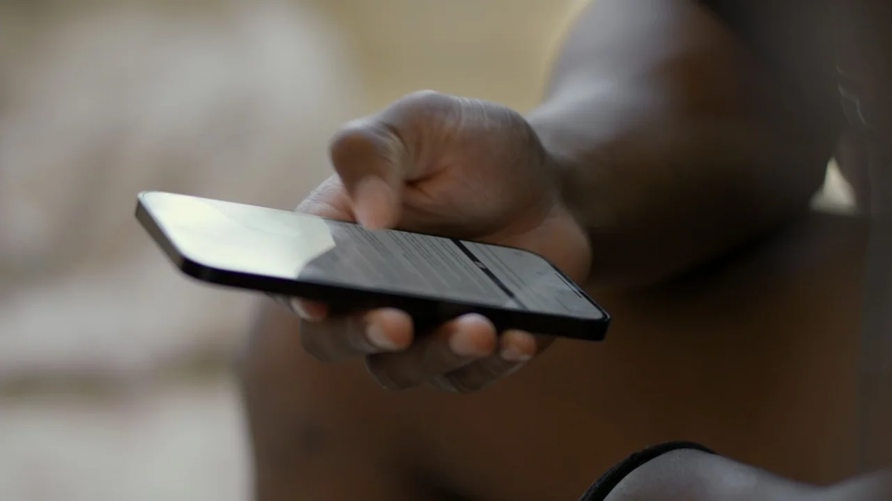 SAQUON (2025) movie still: closeup — A close-up view shows a person's hand holding a dark-rimmed smartphone against a blurred background. The…