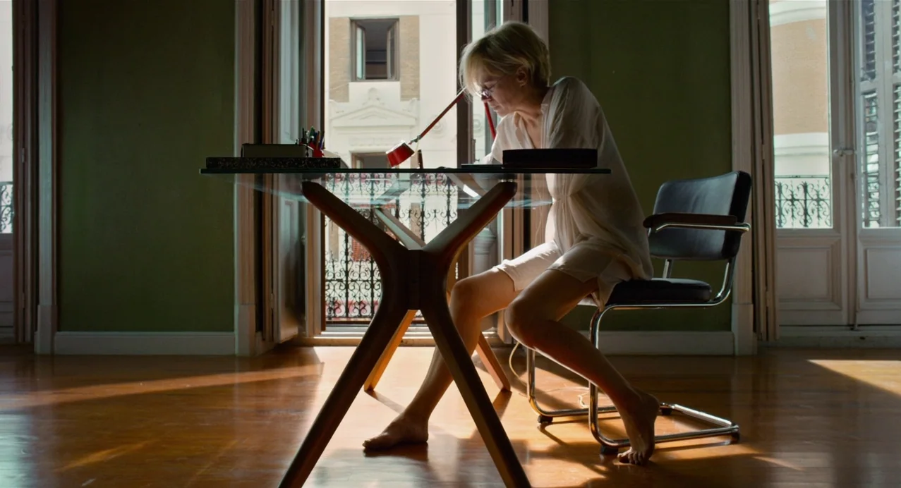 Julieta (2016) movie still: medium — A spacious room features olive green walls and polished wood floors illuminated by sunlight. A woman works at a…