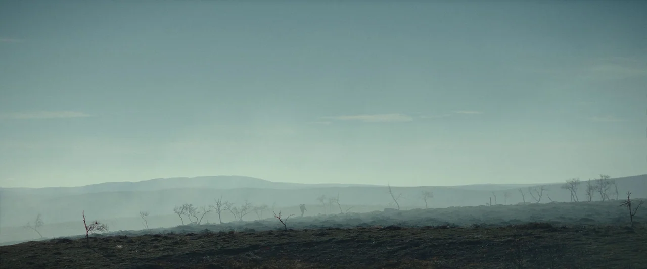 Sisu (2022) movie still: wide — A vast, flat, and misty tundra stretches toward distant rolling hills under a hazy sky. A solitary figure carrying a…