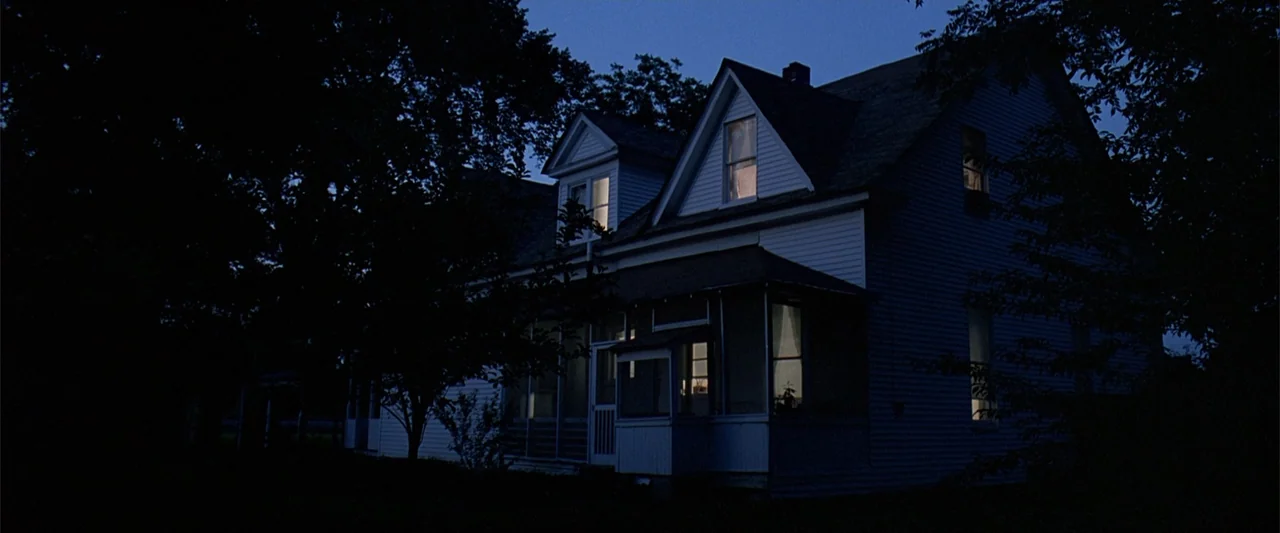 A Family Thing (1996) movie still: establishing — A two-story white farmhouse sits surrounded by dark trees at late blue hour. Several windows on…